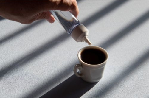 Mão segurando um frasco de adoçante colocando dentro de uma xícara de café para representar o aspartame